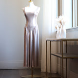 A sleeveless, floor-length satin slip dress in a dusty rose pink color displayed on a mannequin stand in a bright room next to a window.