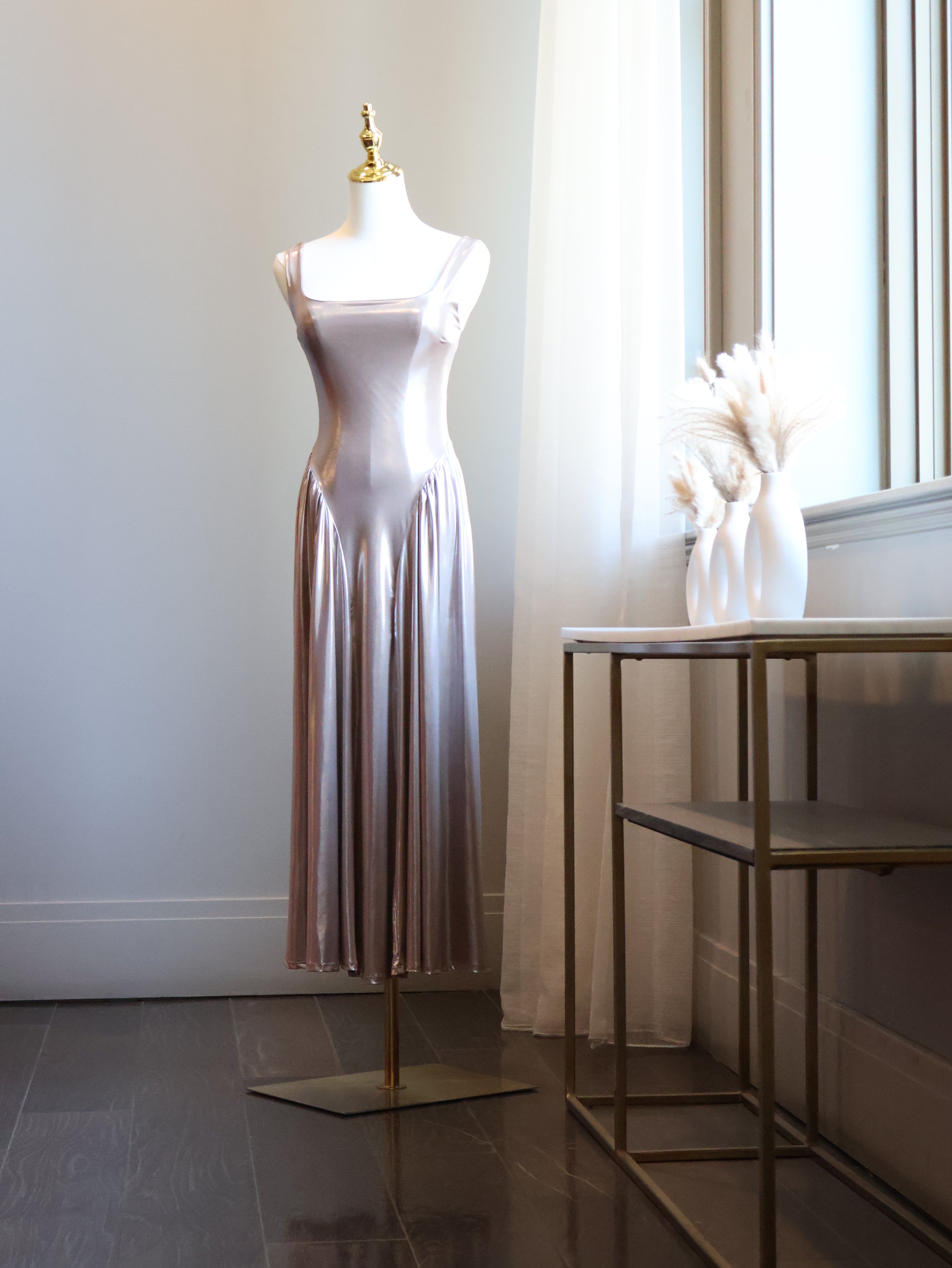 A sleeveless, floor-length satin slip dress in a dusty rose pink color displayed on a mannequin stand in a bright room next to a window.